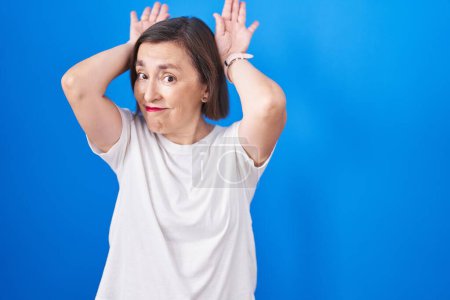 Photo for Middle age hispanic woman standing over blue background doing bunny ears gesture with hands palms looking cynical and skeptical. easter rabbit concept. - Royalty Free Image
