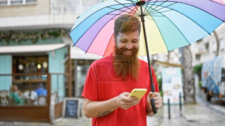 Foto de Joven pelirrojo confiado sonriendo alegremente, sosteniendo un paraguas y usando un teléfono inteligente en una bulliciosa calle de la ciudad. - Imagen libre de derechos