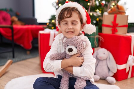 Foto de Adorable niño hispano abrazando oso de peluche sentado en el suelo por el árbol de Navidad en casa - Imagen libre de derechos