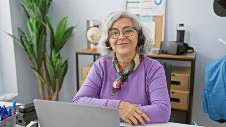 Foto de Mujer madura sonriente con el pelo gris usando gafas y un auricular en un moderno lugar de trabajo de oficina. - Imagen libre de derechos