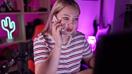 Photo for A surprised young blonde woman talking on the phone in a colorful gaming room at night - Royalty Free Image