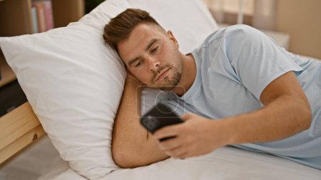 A young hispanic man with a beard lounging in a bedroom, looking at a smartphone while reclining on a pillow.