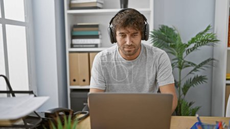 Photo for Focused hispanic man with beard and headphones working on laptop in modern office interior. - Royalty Free Image