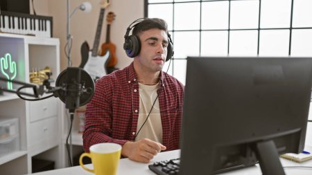 Passionate young hispanic male musician, engrossed in his craft, immersed in melodies at the music studio, using computer and headphones, sipping coffee while serious and focused