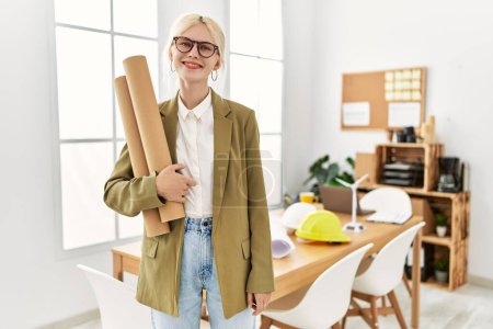Photo for Young blonde woman architect smiling confident holding blueprints at office - Royalty Free Image