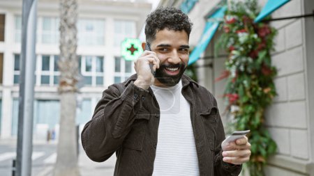 Téléchargez les photos : Bel homme barbu parler sur smartphone tout en tenant la carte de crédit sur la rue de la ville urbaine. - en image libre de droit