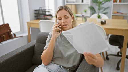 Photo for A focused young woman discusses work on a phone call in a modern office, holding documents. - Royalty Free Image