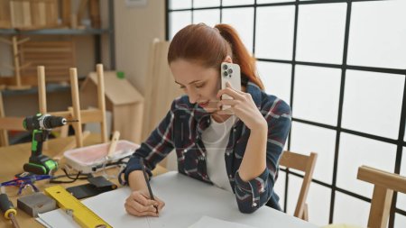 Téléchargez les photos : Une femme concentrée multitâche dans un atelier de menuiserie tout en parlant au téléphone et en écrivant des notes. - en image libre de droit