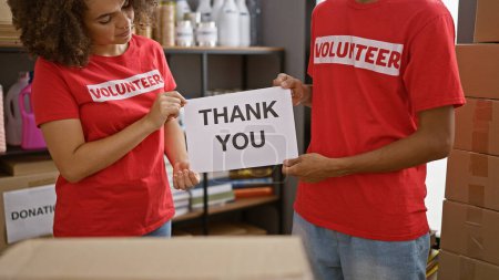 Photo for In the heart of activism, man and woman volunteers at charity center standing united, holding 'thank you' message. young, beautiful community workers in t-shirts, showing gratitude amidst donations. - Royalty Free Image