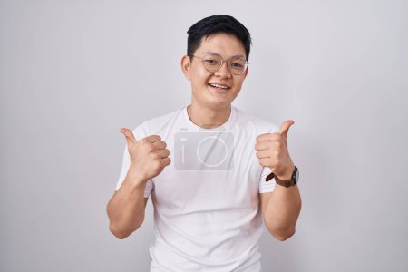 Photo for Young asian man standing over white background success sign doing positive gesture with hand, thumbs up smiling and happy. cheerful expression and winner gesture. - Royalty Free Image