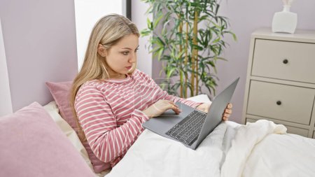 Photo for A focused blonde woman uses a laptop while relaxing on a bed in a cozy bedroom setting. - Royalty Free Image