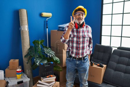 Photo for Cheerful senior man in hardhat and safety glasses, proudly showing the 'two' sign with fingers while joyously pointing up at his new home - Royalty Free Image