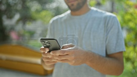 Photo for Handsome young, blond, bearded man using his smartphone for texting conversation in green city park. adult guy immersed in online communication, holding mobile, deep in digital connection. - Royalty Free Image