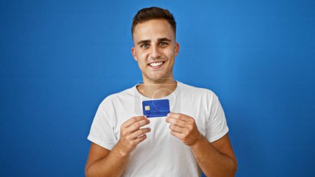Téléchargez les photos : Homme hispanique souriant tenant une carte de crédit sur fond bleu, dépeignant la confiance dans la finance. - en image libre de droit