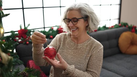 Téléchargez les photos : Femme d'âge moyen avec les cheveux gris souriant décoration confiante arbre de Noël à la maison - en image libre de droit