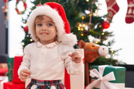 Foto de Adorable chica rubia sonriendo confiada de pie junto al árbol de Navidad en casa - Imagen libre de derechos