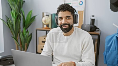 Foto de Un joven alegre hispano con barba que usa auriculares sonríe mientras trabaja en un portátil en un entorno de oficina moderno. - Imagen libre de derechos
