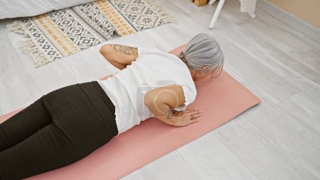 Foto de Entrenamiento de mujer de mediana edad de pelo gris con estiramientos de yoga en la estera, en el entrenamiento en casa para el fortalecimiento saludable de la espalda, expresión emotiva de atleta femenina madura - Imagen libre de derechos