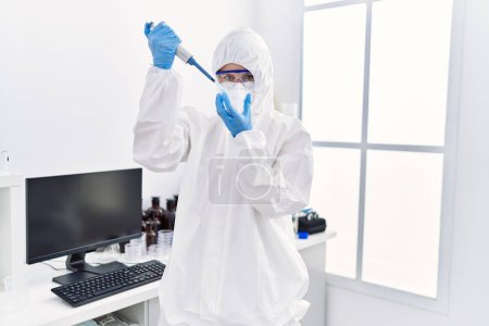 Foto de Joven mujer rubia científica con uniforme de seguridad vertiendo líquido en la muestra en el laboratorio - Imagen libre de derechos