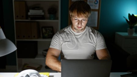 Konzentrierter junger Mann aus dem Kaukasus arbeitet spät am Laptop in einem Homeoffice-Setup und zeigt Professionalität und Engagement.
