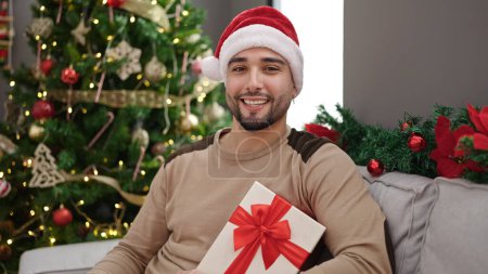 Photo for Young arab man holding gift sitting on sofa by christmas tree at home - Royalty Free Image