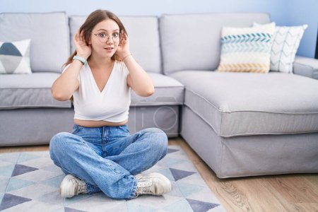 Photo for Young caucasian woman sitting on the floor at the living room trying to hear both hands on ear gesture, curious for gossip. hearing problem, deaf - Royalty Free Image