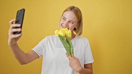 Téléchargez les photos : Jeune femme blonde tenant bouquet de fleurs faisant selfie par smartphone sur fond jaune isolé - en image libre de droit