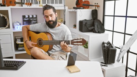 Photo for Handsome bearded hispanic man playing guitar in a modern music studio with instruments. - Royalty Free Image