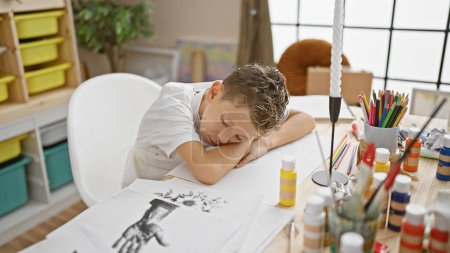 Téléchargez les photos : Adorable garçon artiste blond, appuyé sur la table, dormant après une leçon de peinture fatigante à l'intérieur au studio d'art - en image libre de droit