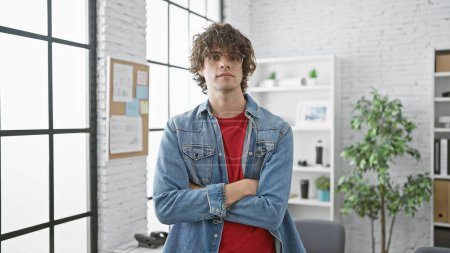 Foto de Un joven con el pelo rizado y una barba se pone de pie brazos cruzados en una oficina moderna brillante, exudando confianza. - Imagen libre de derechos