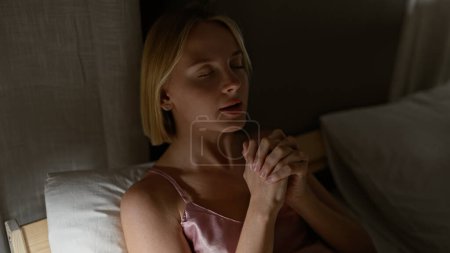 Foto de Mujer rubia joven sentada en la cama rezando en el dormitorio - Imagen libre de derechos