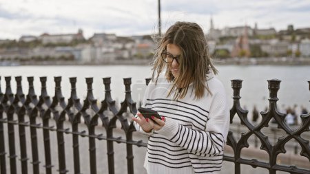 Foto de Una joven usando un teléfono inteligente junto al río Danubio en budapest, con paisaje urbano en el fondo. - Imagen libre de derechos