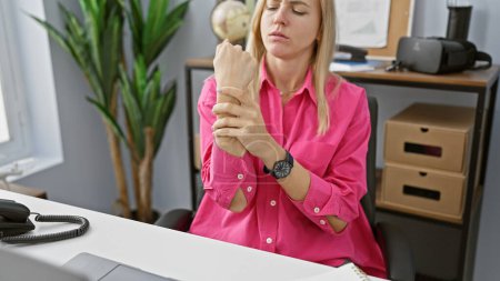 Téléchargez les photos : Une femme blonde en chemise rose ressent une douleur au poignet à son bureau - en image libre de droit
