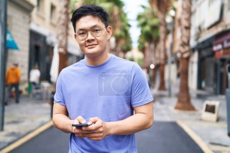 Foto de Joven hombre chino sonriendo confiado usando teléfono inteligente en la calle - Imagen libre de derechos