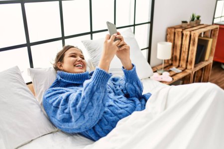 Téléchargez les photos : Jeune belle femme hispanique faire selfie par smartphone couché sur le lit dans la chambre - en image libre de droit
