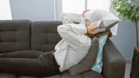 Foto de Mujer rubia estresada acostada en un sofá gris en casa cubriendo la cara con almohada - Imagen libre de derechos