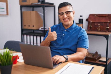 Téléchargez les photos : Jeune homme hispanique travaillant au bureau avec un ordinateur portable faisant des pouces heureux geste avec la main. approuvant l'expression regardant la caméra montrant le succès. - en image libre de droit