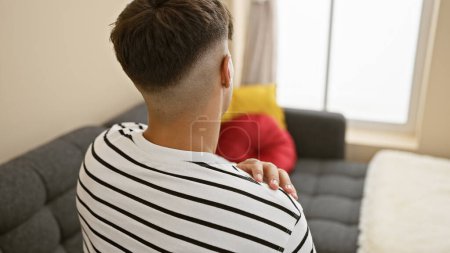 Foto de Hombre hispano joven preocupado sentado en el sofá en casa, sufriendo dolor de espalda grave, una realidad dolorosa en el interior. - Imagen libre de derechos