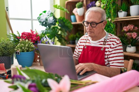 Téléchargez les photos : Moyen Age homme aux cheveux gris fleuriste utilisant un ordinateur portable avec une expression sérieuse à la boutique de fleurs - en image libre de droit