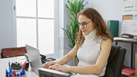 Foto de Mujer hispana enfocada trabajando en una computadora portátil en una moderna configuración de oficina, mostrando productividad y profesionalidad. - Imagen libre de derechos