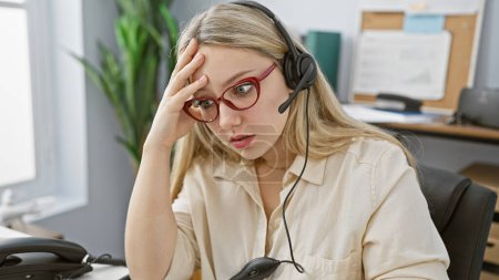 Foto de Una joven rubia estresada con gafas y auriculares en una oficina. - Imagen libre de derechos