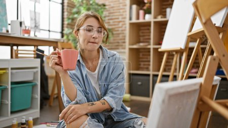 Téléchargez les photos : Jeune femme artiste regardant dessiner tenant tasse de café au studio d'art - en image libre de droit