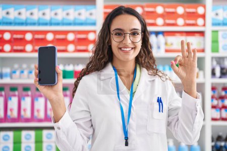 Téléchargez les photos : Jeune femme hispanique travaillant à la pharmacie drugstore montrant écran smartphone souriant regardant vers le côté et regardant loin en pensant. - en image libre de droit