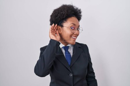 Téléchargez les photos : Belle femme africaine aux cheveux bouclés portant une veste d'affaires et des lunettes souriant main sur oreille en écoutant une rumeur ou des commérages. concept de surdité. - en image libre de droit