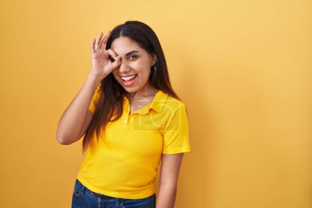 Foto de Mujer árabe joven de pie sobre fondo amarillo haciendo buen gesto con la mano sonriente, ojos mirando a través de los dedos con la cara feliz. - Imagen libre de derechos