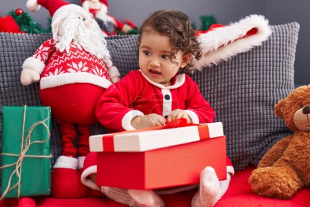 Foto de Adorable niño hispano desempacando regalo de Navidad sentado en un sofá en casa - Imagen libre de derechos