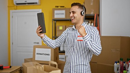 Foto de Un joven alegre con auriculares celebra con un puño en un almacén lleno de cajas de donaciones. - Imagen libre de derechos