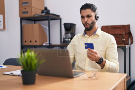 Photo for Hispanic young man wearing call center agent headset doing online shopping scared and amazed with open mouth for surprise, disbelief face - Royalty Free Image