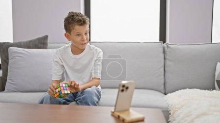 Foto de Adorable chico rubio sentado en el sofá, resolviendo con confianza el rompecabezas del cubo de Rubik, grabando video tutorial casero interior con una expresión linda y sonriente - Imagen libre de derechos
