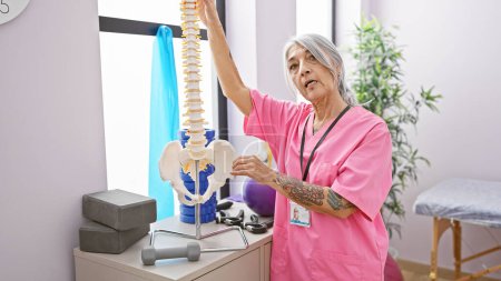Téléchargez les photos : Femme aux cheveux gris sérieuse, physiothérapeute chevronnée, tenant un modèle de colonne vertébrale anatomique, expliquant passionnément la kinésiologie dans une clinique de réadaptation animée - en image libre de droit